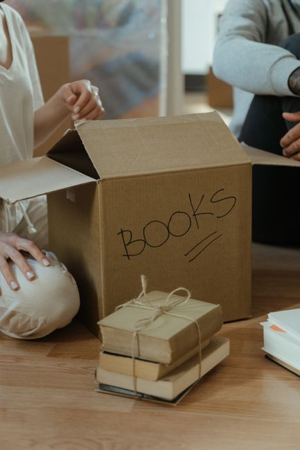 A person dressed in a white shirt is seen loading a small cardboard box wrapped in plastic bubble wrap into a large cardboard moving box labeled 'KITCHEN'. The large box is placed on a patterned tiled